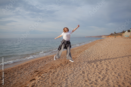 A beautiful middle-aged woman walks on a sandy beach in autumn or spring. A 45-year-old woman is wearing jeans, a beige jacket and a plaid shirt. The active model jumps