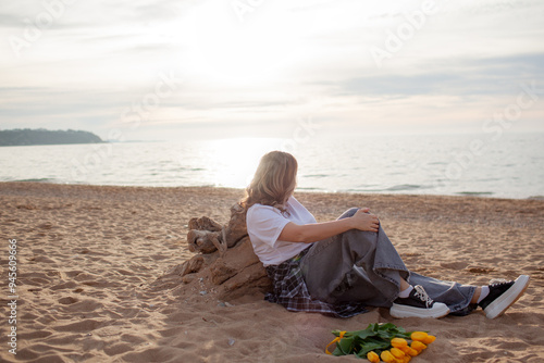 A beautiful middle-aged woman walks on a sandy beach in spring and holds a bouquet of tulips. A 45-year-old woman is wearing jeans, a beige jacket and a plaid shirt.