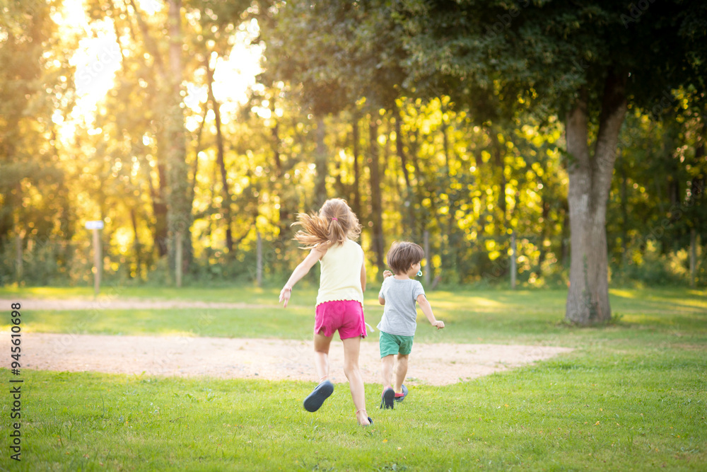 Two kids, boy and girl, back view, siblings, running on the grass at the park at sunset. Background with copy space. Two kids, boy and girl, back view, siblings, running on the grass at the park at sunset. Background with copy space.