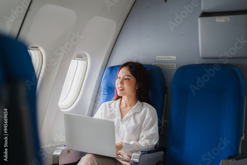 Businesswoman on Airplane: A young woman in a white shirt gazes out the window of an airplane, working on her laptop. The photo evokes a sense of travel, ambition, and the pursuit of dreams.