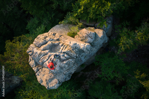 Aerial view of climber standing atop rock formation at Dovbush Rocks in Carpathian mountains, Ukraine. Sun sets, casting warm glow over lush, green forest, distant hills, creating breathtaking view. photo