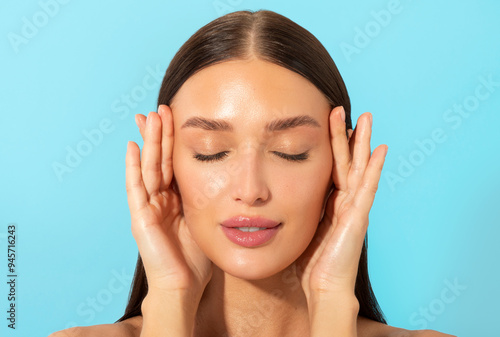 Close-up portrait of attractive European woman touching temples, showing her perfect glowing clean skin, posing isolated over blue background