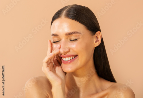 Portrait of young European brunette woman smiling with eyes closed, touching her face gently over beige studio backdrop, showing natural beauty