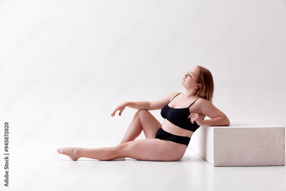 Calm and relaxed young beautiful woman with overweight body shape posing on floor in underwear isolated on white studio background. Concept of female beauty, body positivity, care, acceptance