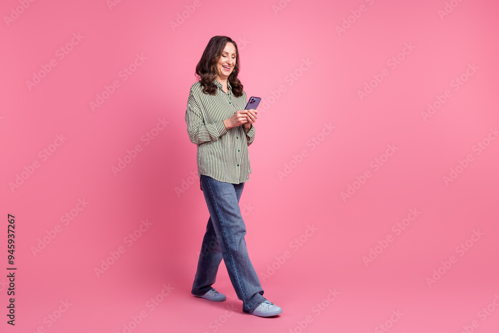 Full size photo of nice aged woman walk use smart phone wear shirt isolated on pink color background Full size photo of nice aged woman walk use smart phone wear shirt isolated on pink color background