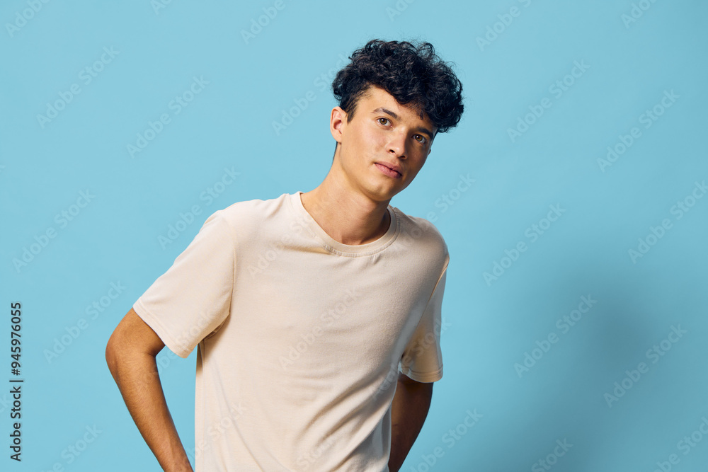 Young man with curly hair wearing a light beige t shirt against a solid blue background, exhibiting a casual and stylish pose, ideal for fashion and lifestyle themes Young man with curly hair wearing a light beige t shirt against a solid blue background, exhibiting a casual and stylish pose, ideal for fashion and lifestyle themes
