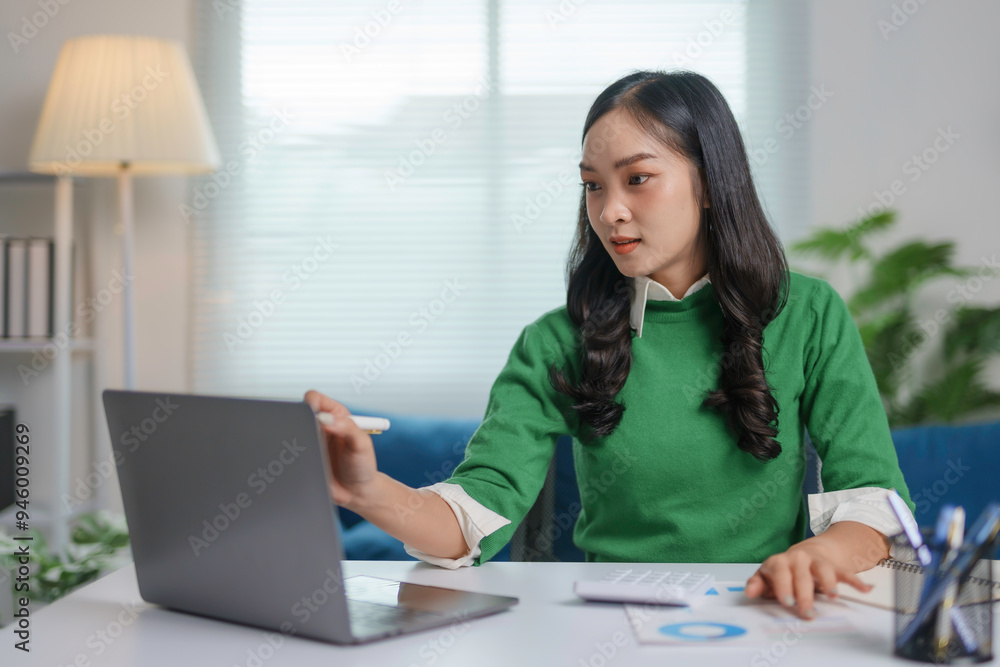 Asian accountant is analyzing financial data on her laptop, pointing at the screen with a pen while working from her home office Asian accountant is analyzing financial data on her laptop, pointing at the screen with a pen while working from her home office