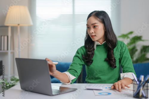 Asian accountant is analyzing financial data on her laptop, pointing at the screen with a pen while working from her home office