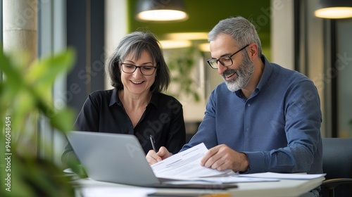 Two happy busy middle aged professionals man and woman business leaders partners checking document reading financial report talking working together on laptop computer.