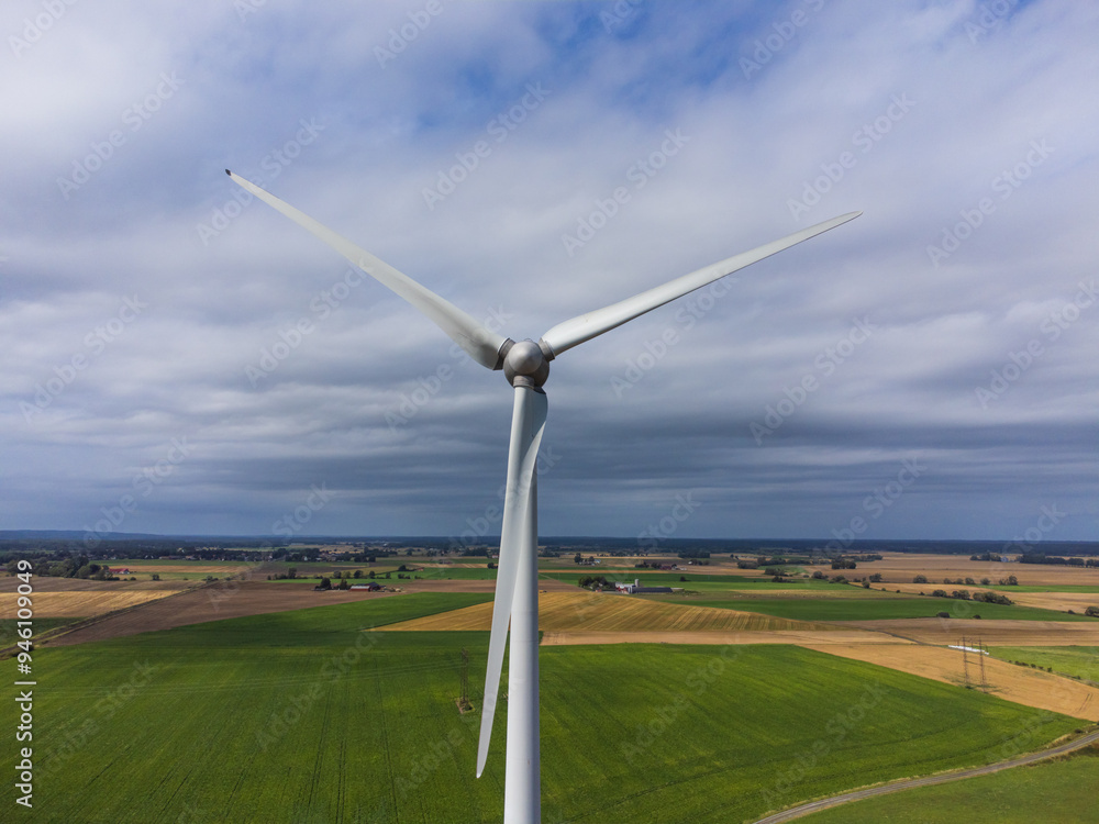 Aerial front view of a wind turbine in the wind. Sweden, summer, green and yellow fields and farmland. Cloudy with sunrays illuminating the wind turbine. Aerial front view of a wind turbine in the wind. Sweden, summer, green and yellow fields and farmland. Cloudy with sunrays illuminating the wind turbine.