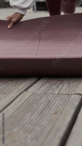 Vertical slowmo shot of senior Caucasian woman in activewear rolling out yoga mat on wooden flooring in piblic park for training outdoors photo