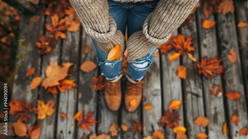 A close-up of an adult wearing a cozy knit sweater, paired with jeans and boots, standing on a rustic wooden porch with autumn leaves scattered around