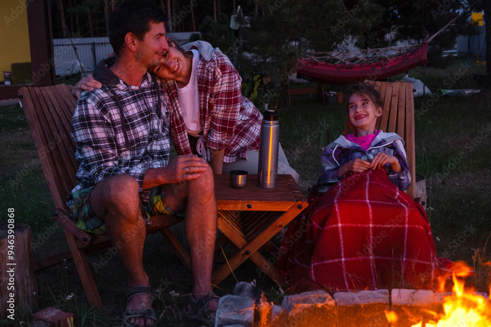 Fototapeta premium Family is sitting by the outdoor campfire in the courtyard of the house on camping chairs, drink tea from a thermos, warm up and chat by the hearth
