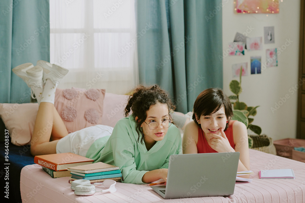 Two young women lying on bed in cozy bedroom, studying together using a laptop, with books, pillows, and decorations around them creating a warm atmosphere Two young women lying on bed in cozy bedroom, studying together using a laptop, with books, pillows, and decorations around them creating a warm atmosphere