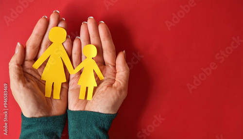 Hands holding a family recreated in yellow paper, consisting of a mother and daughter, on a red background. The person holding the concept of family unity is wearing a green sweater