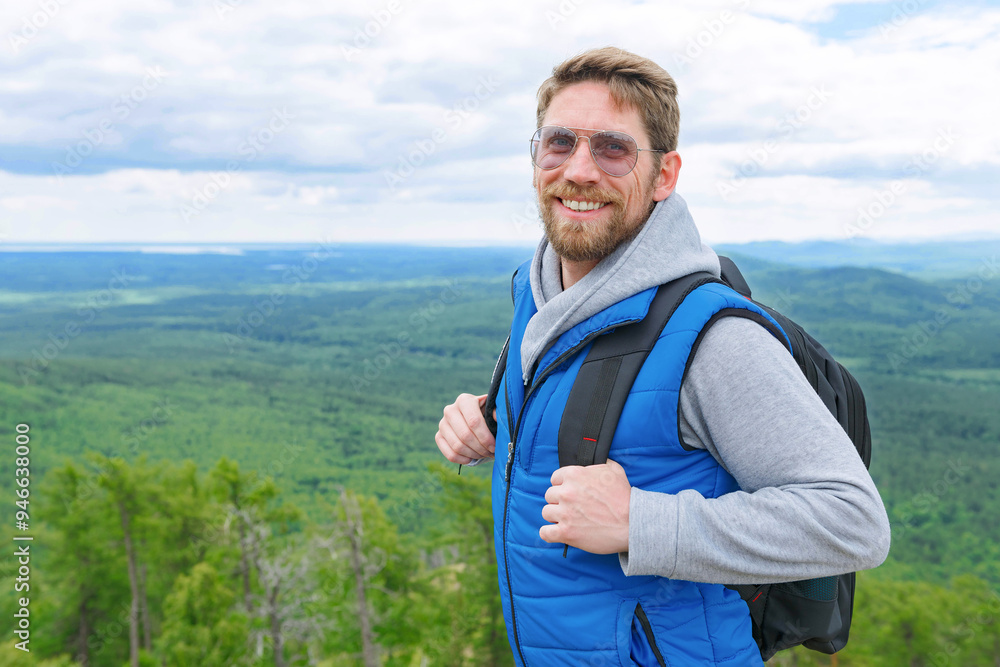 A man in a blue tank top and grey turtleneck smiles at the camera on a mountain top with a backpack. A man in a blue tank top and grey turtleneck smiles at the camera on a mountain top with a backpack.