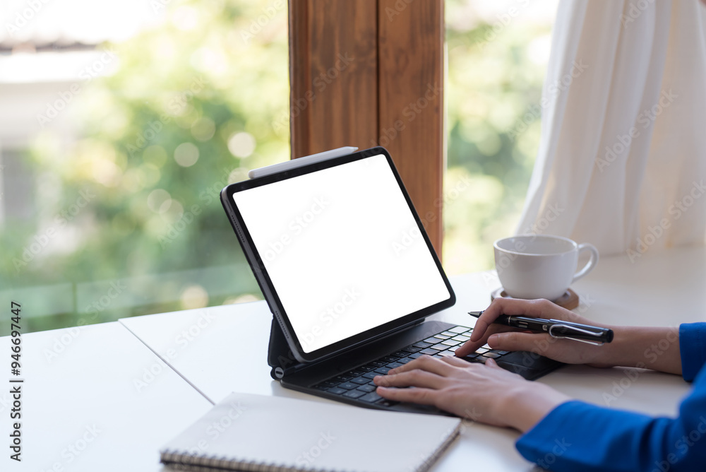 Fototapeta premium Businesswoman working from her home office with a tablet, typing and connecting online in a bright, organized space with a nature view. Ideal for remote work and freelance opportunities