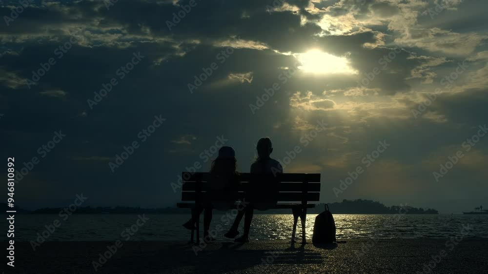 Two girls on bench by evening river. A view of two little girls silhouette relax on the bench and admire the river during nightfall. Two girls on bench by evening river. A view of two little girls silhouette relax on the bench and admire the river during nightfall.
