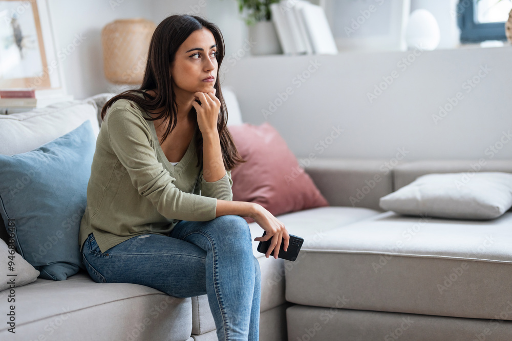Obraz premium Worried young woman using her mobile phone while thinking about problems sitting on couch in the living room at home.