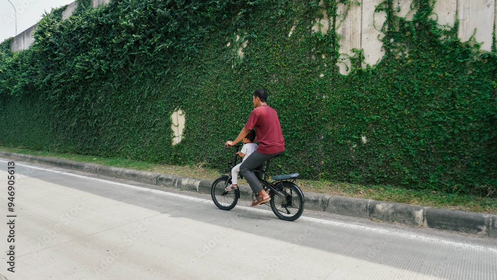 Obraz premium A father and son riding a bicycle on a quiet road, flanked by a tall green ivy wall. A serene urban scene capturing familial joy