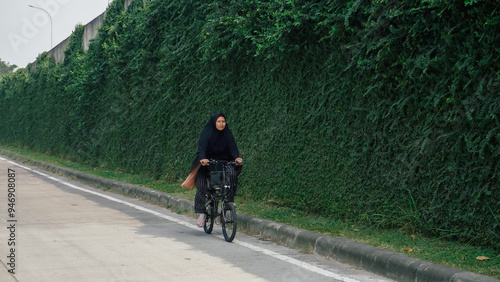 A woman dressed in black hijab cycles next to a wall thick with ivy, highlighting a balance between modern transportation and nature photo