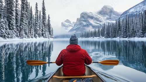 A kayaker paddles through a snowy landscape on a serene lake surrounded by towering mountains and dense evergreen forests in winter