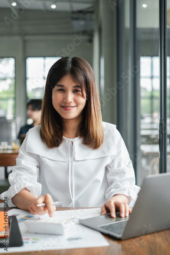 Smiling young woman in a white blouse working on a laptop in a bright, modern office setting with large windows.