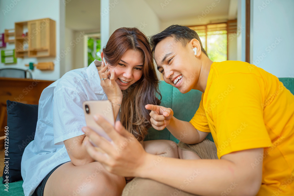 Indonesian couples are sitting on a couch, looking at a cell phone while video calling, listening to music, or watching a movie Indonesian couples are sitting on a couch, looking at a cell phone while video calling, listening to music, or watching a movie