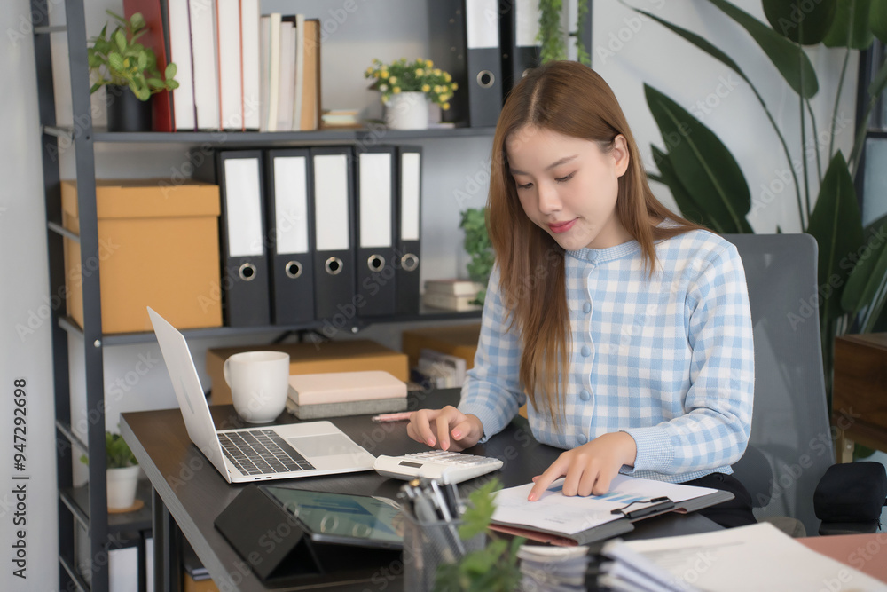 An Asian female businesswoman is sitting at her desk, working on multiple tasks. She is focused on her documents, calculator, and laptop, fully immersed in a modern, organized workspace An Asian female businesswoman is sitting at her desk, working on multiple tasks. She is focused on her documents, calculator, and laptop, fully immersed in a modern, organized workspace