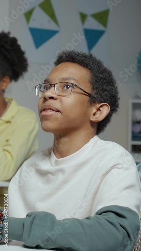 Vertical medium close-up of teenage African American boy sitting at desk in classroom having conversation witth offscreen teacher during lesson photo