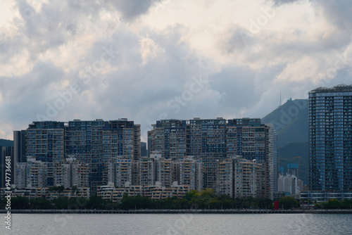 Photography High-rise residential buildings along the waterfront in Hong Kong under a dramatic cloudy sky with green mountains in the background