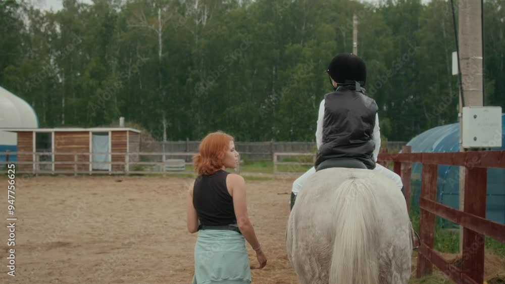 Back view of unrecognizable child wearing white breeches, black equestrian helmet and paddock boots sitting on saddle on horseback during outdoor horse riding lesson with female trainer Back view of unrecognizable child wearing white breeches, black equestrian helmet and paddock boots sitting on saddle on horseback during outdoor horse riding lesson with female trainer