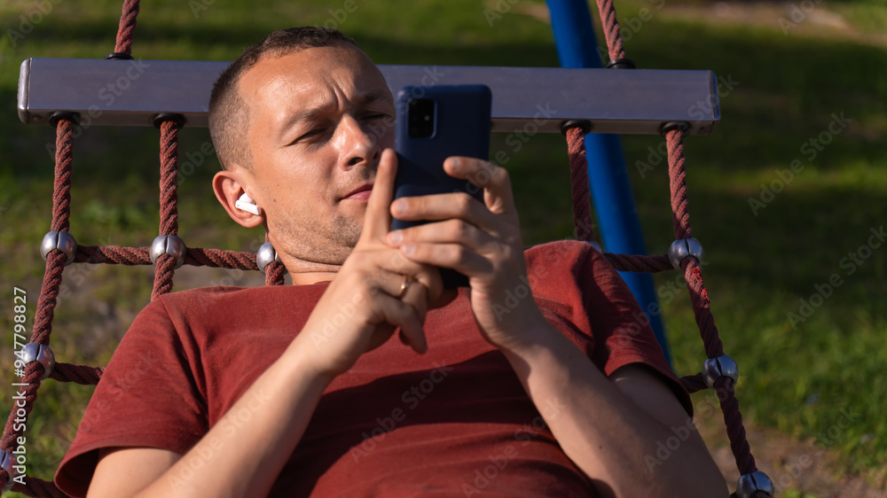 Obraz premium Portrait of a man looking at a mobile phone lying in a hammock and listening to music through white wireless headphones in the park.