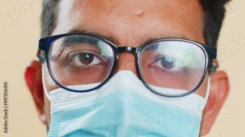 Extreme close up face portrait of Indian young doctor cardiologist man lab worker in glasses looking at camera. Arabian apothecary pharmacy guy in medical protective mask isolated on beige background photo