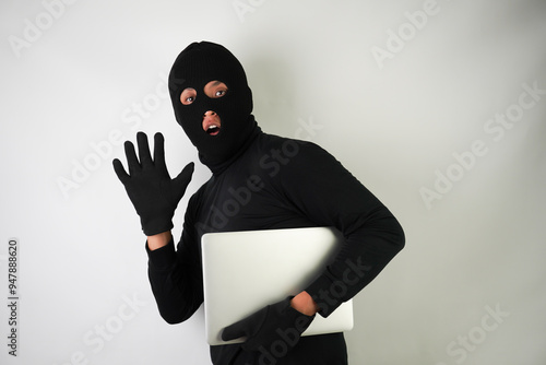 Young Adult black masked robber is holding silver colored laptop. Running, fleeing and escaping pose. Concept for hacking, scamming, phishing.