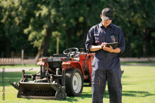 In cap, holding digital tablet. Man is with utility tractor with grass cutter and aerator equipment on the field