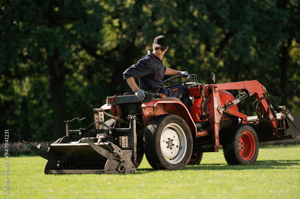 Fototapeta premium Sitting and driving. Man is with utility tractor with grass cutter and aerator equipment on the field