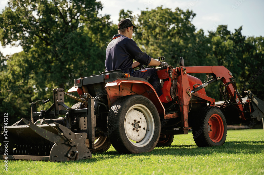 Fototapeta premium Sitting and driving. Man is with utility tractor with grass cutter and aerator equipment on the field