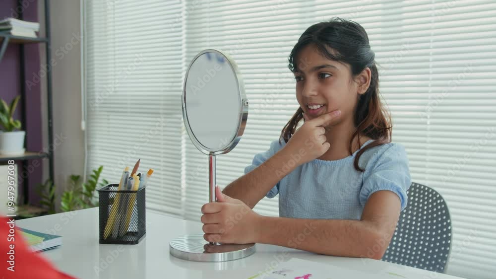Medium shot of teenage Indian schoolgirl pronouncing sounds by mirror when sitting at table in classroom during speech therapy session with tutor Medium shot of teenage Indian schoolgirl pronouncing sounds by mirror when sitting at table in classroom during speech therapy session with tutor