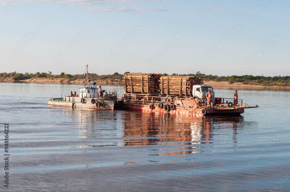 River ferry carries a truck loaded with logs on the Northern Dvina near Krasnoborsk, Russia. Summer time, sunset River ferry carries a truck loaded with logs on the Northern Dvina near Krasnoborsk, Russia. Summer time, sunset