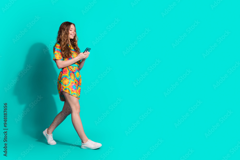 Full body photo of attractive teen woman hold device walking dressed stylish colorful clothes isolated on cyan color background Full body photo of attractive teen woman hold device walking dressed stylish colorful clothes isolated on cyan color background