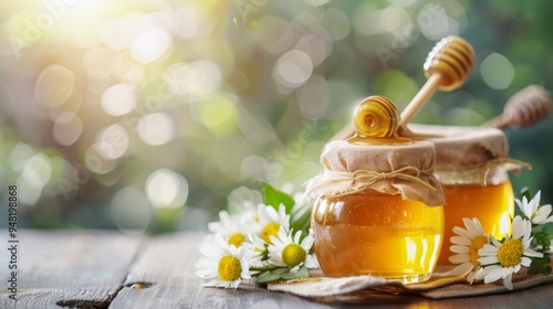 Two jars of honey with daisy decorations and a wooden spoon on a wooden surface. Background hints at a garden with bright lighting.