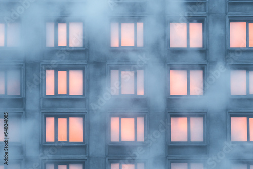 A building with many windows and a foggy atmosphere. The windows are lit up with a warm orange glow