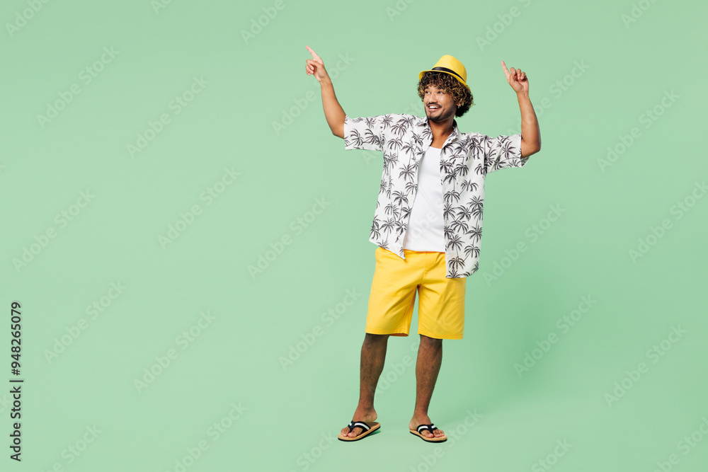 Full body young smiling fun Indian man wear beach shirt hat rest near hotel pool point index finger aside on area dancing isolated on plain green color background. Summer vacation sea sun tan concept. Full body young smiling fun Indian man wear beach shirt hat rest near hotel pool point index finger aside on area dancing isolated on plain green color background. Summer vacation sea sun tan concept.