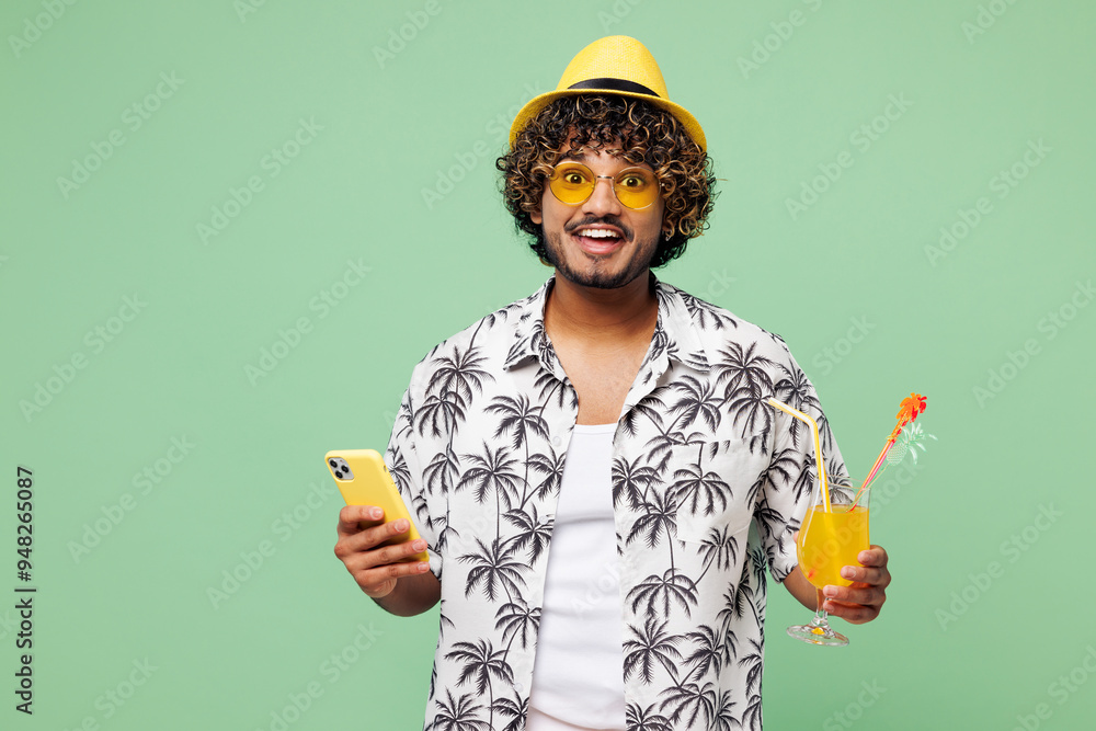 Young Indian man wear beach shirt hat sunglasses drink orange citrus cocktail use mobile cell phone rest near hotel pool isolated on plain green color background. Summer vacation sea sun tan concept. Young Indian man wear beach shirt hat sunglasses drink orange citrus cocktail use mobile cell phone rest near hotel pool isolated on plain green color background. Summer vacation sea sun tan concept.