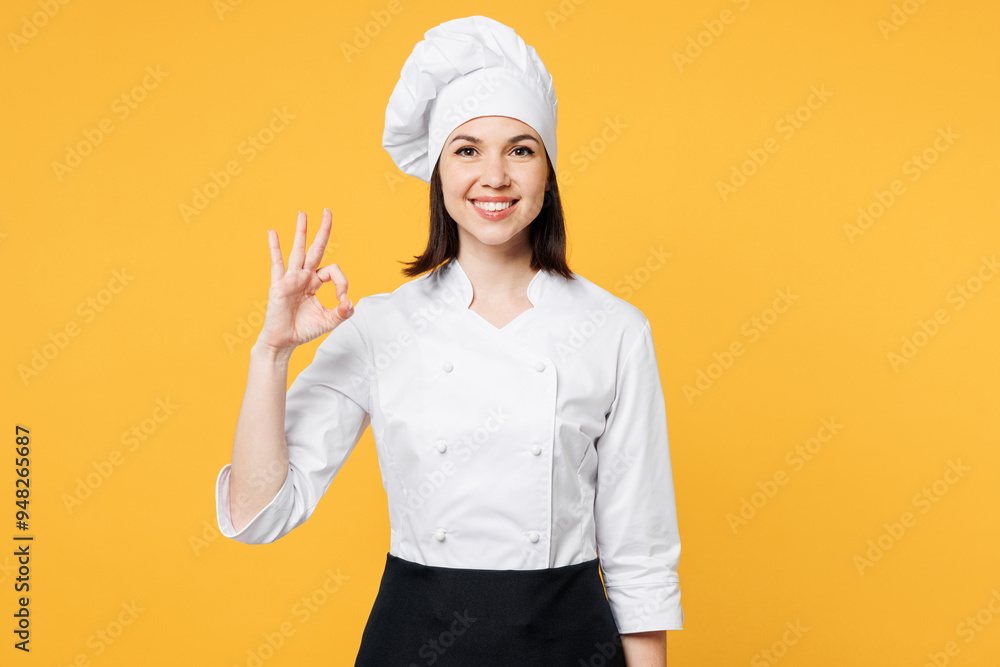 Young smiling happy satisfied chef cook baker woman she wear white shirt black apron uniform toque chefs hat showing okay ok gesture isolated on plain yellow background studio. Cooking food concept. Young smiling happy satisfied chef cook baker woman she wear white shirt black apron uniform toque chefs hat showing okay ok gesture isolated on plain yellow background studio. Cooking food concept.