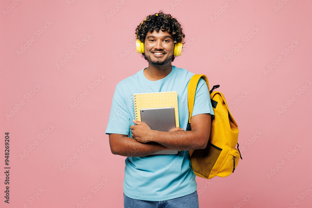 Young happy smart Indian boy student wear blue t-shirt casual clothes backpack bag hold books listen to music in headphones isolated on plain pink background. High school university college concept. Young happy smart Indian boy student wear blue t-shirt casual clothes backpack bag hold books listen to music in headphones isolated on plain pink background. High school university college concept.