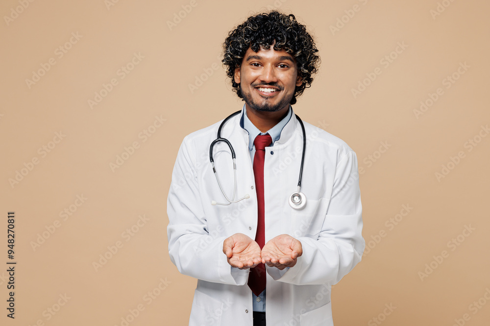 Young Indian doctor man wears white medical gown suit work in hospital clinic office hold empty hands palms pov give something isolated on plain beige background studio. Healthcare medicine concept. Young Indian doctor man wears white medical gown suit work in hospital clinic office hold empty hands palms pov give something isolated on plain beige background studio. Healthcare medicine concept.