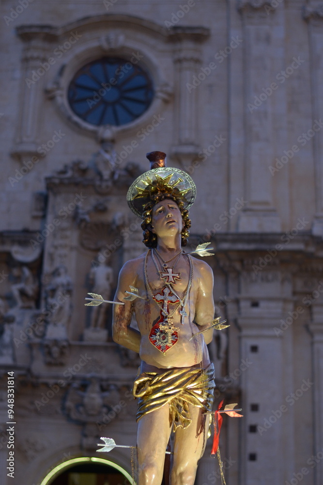 Obraz premium Saint Sebastian Pierced by Arrows on Processional Vara with Illuminated Baroque Church in the Background. Solemn Night Procession with the Statue of Saint Sebastian of Ferla.