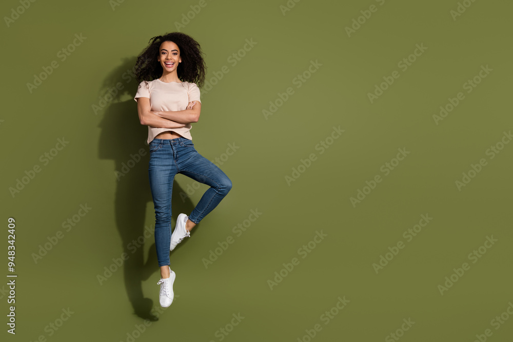 Full size photo of cute girl dressed beige t-shirt jeans jumping arms folded near logo empty space isolated on khaki color background Full size photo of cute girl dressed beige t-shirt jeans jumping arms folded near logo empty space isolated on khaki color background